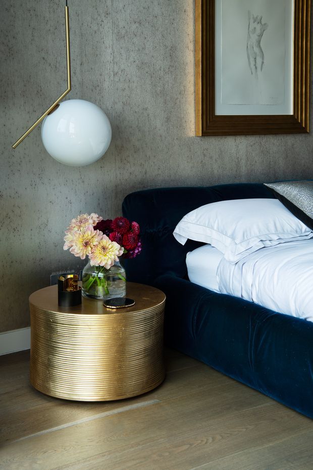 A close-up bedroom scene featuring a dark navy upholstered bed with crisp white bedding, a ribbed cylindrical brass-gold bedside table, a spherical wall lamp with a slim brass arm, textured grey wall finish, timber flooring, and framed artwork. A vase of flowers and small decorative objects sit on the bedside table.
