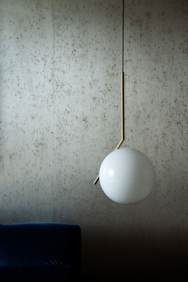 A moody interior detail showing a white spherical pendant light suspended from a slender brass arm against a textured grey concrete wall. The corner of a dark navy upholstered sofa is visible at the lower left, adding contrast to the minimalist composition.