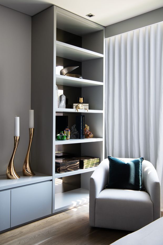 A refined living room corner featuring a built-in grey open shelving unit with integrated lower cabinetry, styled with books and decorative objects. A sculptural white upholstered armchair with a dark teal cushion sits beside full-height sheer white curtains, while metallic gold candleholders add a luxurious accent. Timber flooring and strong natural light complete the contemporary interior.