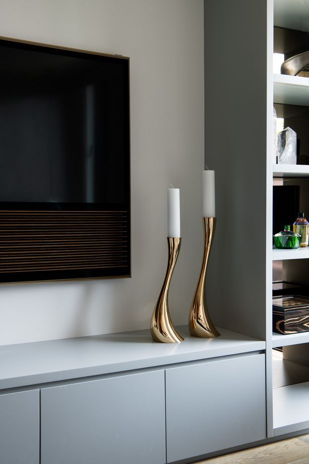 Close-up of a refined contemporary living room media wall, featuring a large black television set within dark horizontal slatted joinery, pale grey built-in cabinetry and shelving, and two tall sculptural gold candleholders. Decorative objects and books are displayed on the open shelving to the right, with timber flooring visible at the bottom.