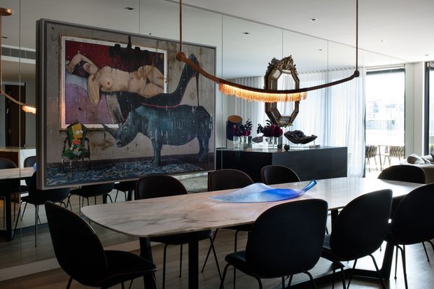 Contemporary dining area with a large sculptural marble dining table surrounded by dark upholstered chairs. Full-height glazing and mirrored surfaces create a bright, open setting, while a large framed artwork, suspended sculptural installation, dark sideboard, and decorative objects add bold eclectic character.