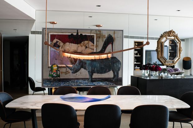A sophisticated dining area with a long pale stone dining table surrounded by dark upholstered chairs. A large framed rhinoceros artwork forms a dramatic focal point behind the table, complemented by sculptural pendant lighting, a black sideboard, a decorative ornate mirror, flowers, and built-in shelving. The space has a refined, eclectic contemporary character with a neutral palette and dark accents.