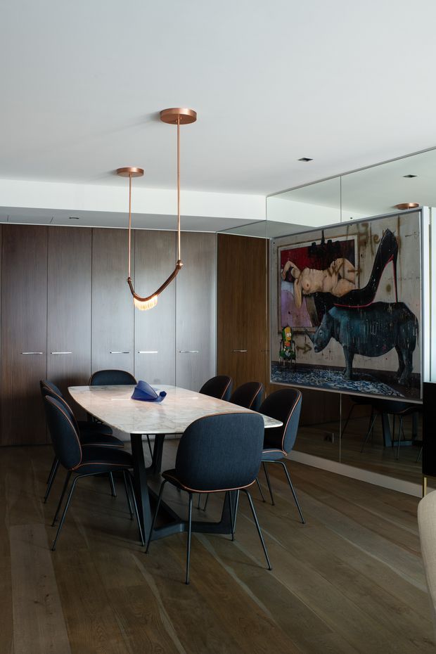 A sophisticated dining room with a large rectangular marble dining table surrounded by dark upholstered chairs. Floor-to-ceiling dark timber cabinetry lines the rear wall, while a full-height mirror reflects the room and artwork. A sculptural copper pendant light hangs above the table, with timber flooring and a large colourful artwork adding visual interest.