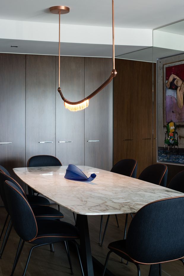 A sophisticated dining room with a large veined stone dining table surrounded by dark upholstered chairs with copper-toned trim. Floor-to-ceiling dark timber cabinetry lines the rear wall, while a sculptural copper pendant light hangs above the table. A mirrored wall and a framed artwork add visual depth and character.