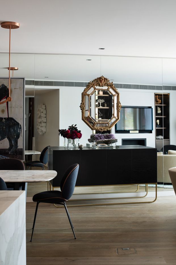 A sophisticated open-plan dining and living interior with a black built-in sideboard or media cabinet, timber flooring, white cabinetry, and an ornate gilded mirror as a focal point. A marble-topped dining table and dark upholstered chairs occupy the foreground, while a wall-mounted television, display shelving, sculptural lighting, and floral arrangements add detail. The room has a polished, high-contrast contemporary aesthetic with classic decorative accents.