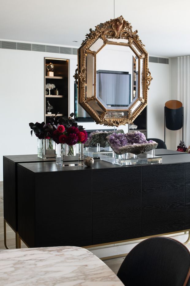 A refined contemporary dining and living space featuring a large black timber sideboard with brass-toned legs, an ornate antique-style gilt mirror, curated floral and mineral displays, a black-and-brass floor lamp, and a marble dining table with dark upholstered seating. White walls, built-in shelving, concealed air-conditioning grilles, and glazing create a bright, polished interior.