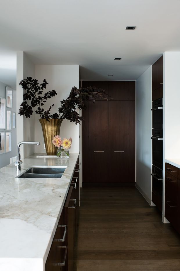 A refined galley-style kitchen with a long white marble island and integrated double sink, dark timber cabinetry, built-in black appliances, and dark timber flooring. Large windows bring in natural light, while a brass-toned vase with foliage and flowers adds a decorative focal point.