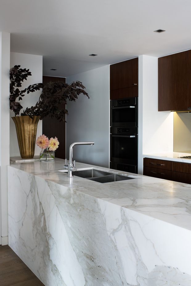 A refined contemporary kitchen centred on a large waterfall island clad in prominently veined white natural stone. The island includes an undermount double sink with a brushed metal gooseneck tap. Dark timber cabinetry and integrated black ovens contrast with white walls and a pale stone benchtop, while under-cabinet lighting illuminates the splashback. A tall decorative vase and flowers sit at the rear of the island.