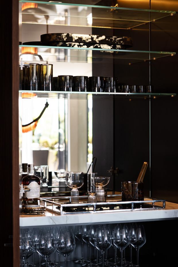 A sophisticated built-in bar cabinet with glossy black cabinetry, mirrored backing, and multiple glass shelves. The display contains black and clear tumblers, wine glasses, decanters, cocktail glasses, trays, and a liquor bottle. A white marble benchtop provides contrast, while integrated warm lighting and reflective surfaces create a dramatic, luxurious atmosphere.