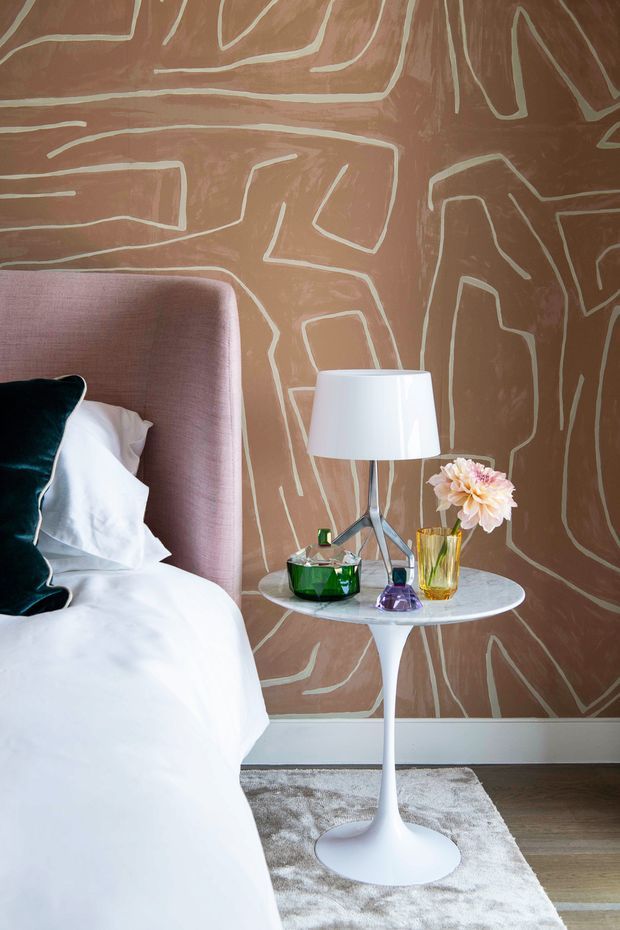 A stylish bedroom corner featuring a tall upholstered blush-pink headboard, white bedding, and a dark teal accent cushion. A white pedestal side table holds a modern lamp, green glass bowl, purple ornament, and a small flower arrangement. The room is defined by a warm brown feature wall with an abstract cream line pattern, light timber flooring, and a pale textured rug.