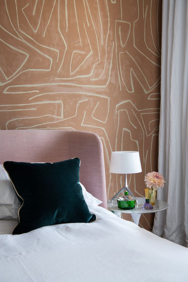 A close-up of a stylish bedroom corner featuring a blush-pink upholstered bedhead, white bedding, a deep teal velvet cushion, and a round side table with a white lamp, green glass vessel, and flowers. The room has a warm terracotta-brown feature wall with an abstract pale linear pattern, alongside full-length white curtains.