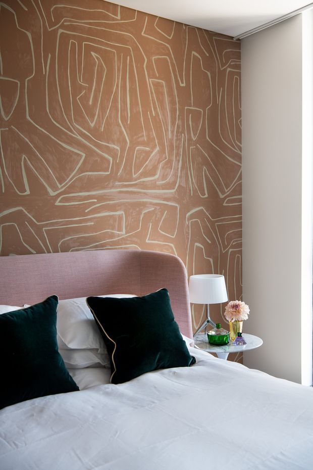 A softly lit bedroom featuring a large upholstered blush-pink bedhead, white bedding, and deep teal velvet cushions. The main feature is a warm terracotta-brown wall with an irregular pale line-art pattern, complemented by a small round bedside table, white lamp, and decorative accessories.