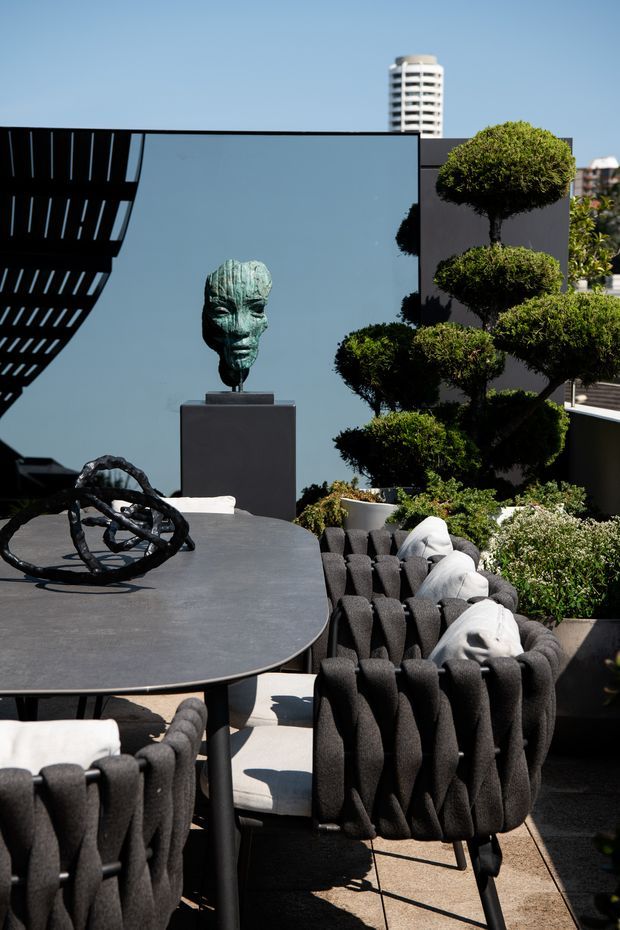 A sophisticated rooftop terrace or outdoor dining area with a dark rounded-edge dining table, sculptural woven charcoal armchairs, a contemporary abstract table sculpture, and a bronze-toned bust on a pedestal. Carefully clipped topiary trees and layered planter greenery frame the space, with a modern urban skyline visible beyond. The scene has strong sunlight, crisp shadows, and a refined contemporary aesthetic.