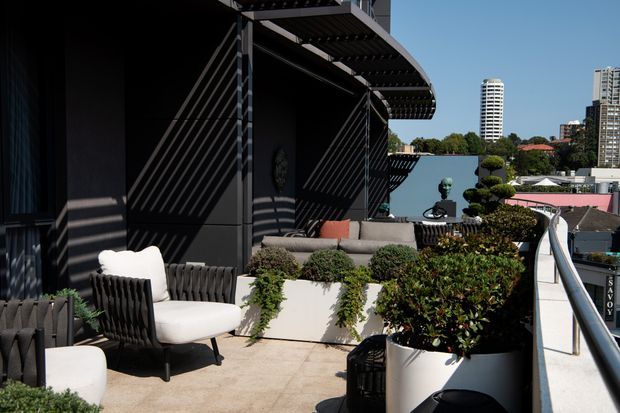 A sophisticated rooftop terrace with dark architectural screening and a curved pergola overhead. The outdoor lounge includes upholstered chairs, a grey sofa with accent cushions, sculptural planting in large white planters, and a projecting seating or garden platform. A city skyline is visible beyond the terrace railing.