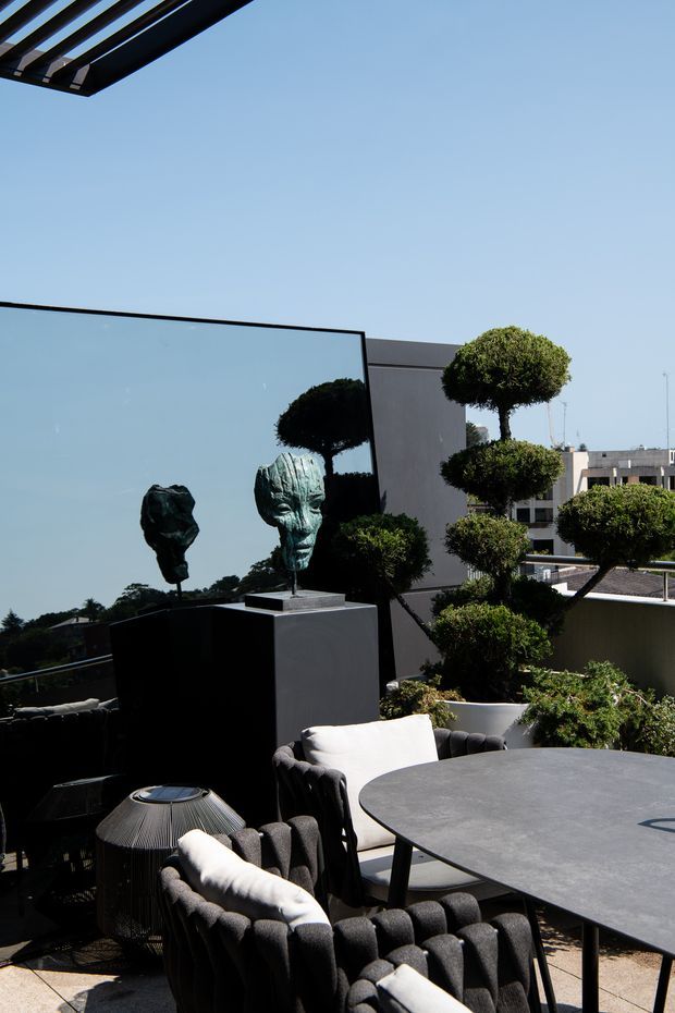 A sophisticated rooftop terrace with a dark round dining table, sculptural upholstered outdoor chairs, a woven side table, topiary plants, and contemporary bust sculptures. A large frameless glass balustrade and adjacent modern building forms frame the open urban setting, with a partial overhead pergola casting shade.