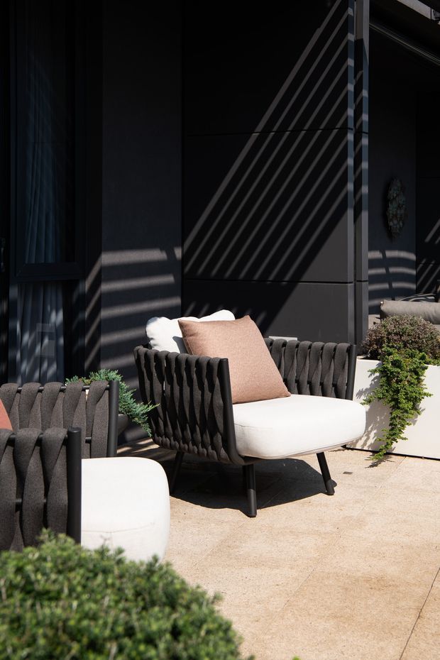 A moody, contemporary outdoor seating area with sculptural black lounge chairs, cream cushions, and a blush accent pillow arranged against a dark paneled facade. Strong diagonal shadows fall across the wall and chairs, while clipped green shrubs in planters soften the space. The area appears to be a refined patio or terrace.