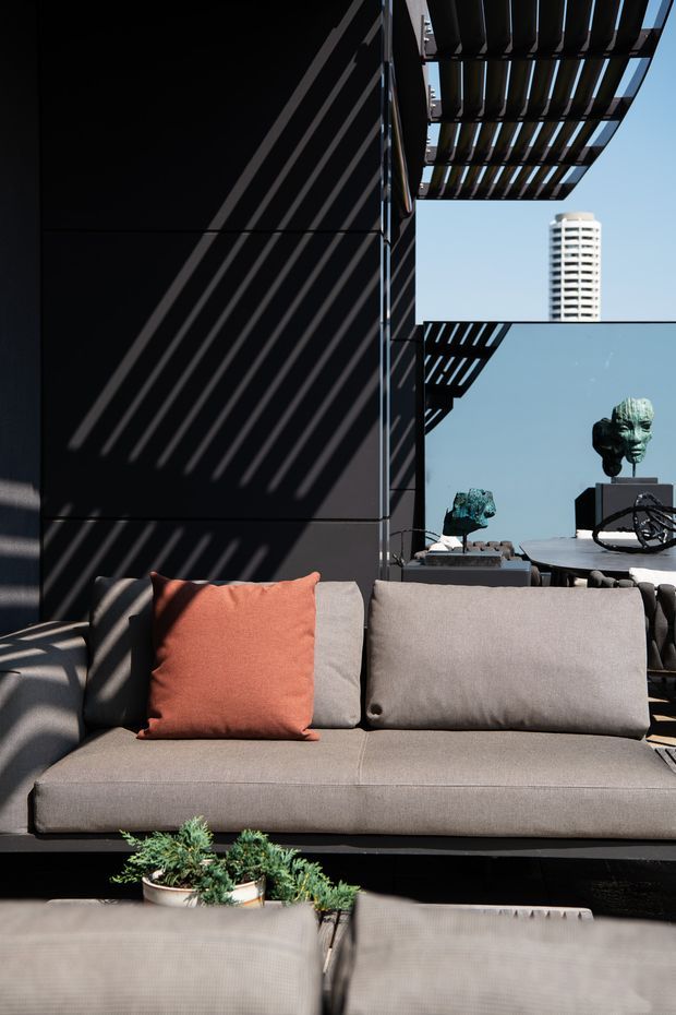 A contemporary outdoor lounge terrace with a large grey upholstered sectional sofa, a terracotta accent cushion, and low planters in the foreground. Dark architectural wall panels and overhead slatted screening cast strong diagonal shadows across the seating area. Sculptural artworks and a city high-rise are visible beyond the terrace, creating a refined urban rooftop atmosphere.