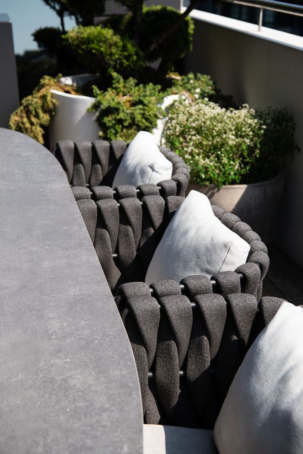 Sunlit outdoor dining area with a long dark grey stone or concrete table and several sculptural woven upholstered dining chairs with white cushions. Curved rendered planter walls and dense greenery create a landscaped terrace setting.