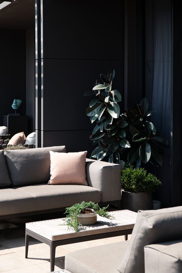 A sophisticated outdoor lounge area with a grey upholstered sofa and armchair arranged around a low rectangular coffee table. The space features dark vertical architectural wall panels, large glazed openings with sheer curtains, and substantial potted greenery. Strong sunlight creates a high-contrast, intimate atmosphere with muted neutral furnishings and dark architectural surfaces.