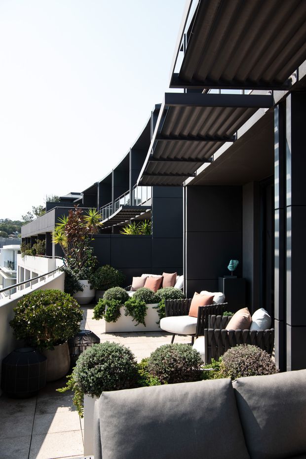 A contemporary outdoor terrace or balcony with a series of covered seating areas along a dark-clad building facade. The space features black metal framing, corrugated overhead awnings, pale tiled paving, upholstered lounge chairs and sofas, and abundant potted shrubs and ornamental plants. The terrace appears elevated, with railings and neighboring balconies visible to the side.