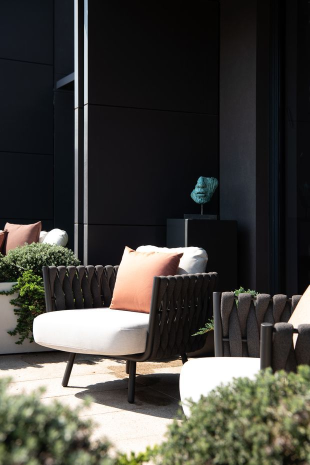 A sophisticated outdoor seating area beside a dark, near-black architectural facade. The scene features woven charcoal lounge chairs with thick white cushions and peach accent pillows, arranged among clipped green shrubs and potted plants on pale paving. A small sculptural object sits on a dark pedestal against the wall. Strong sunlight creates a high-contrast, refined courtyard or terrace atmosphere.