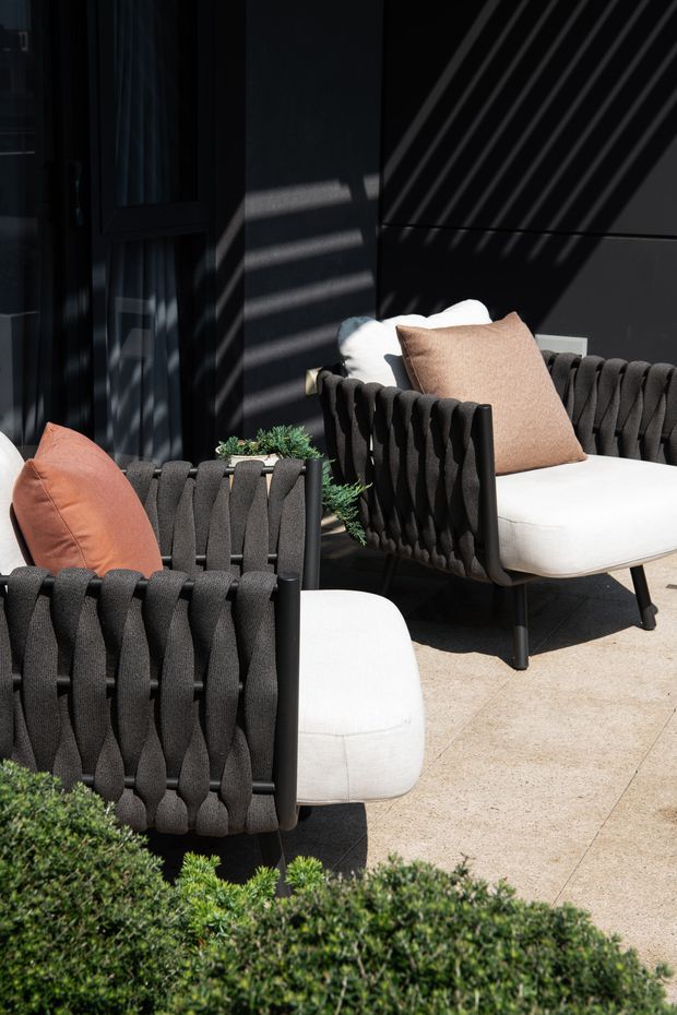 A sunlit outdoor lounge area with two sculptural black woven armchairs, white upholstered cushions, and rust and taupe throw pillows. The seating is arranged on a pale stone or concrete-paved terrace beside a dark exterior wall and glazed door, with clipped greenery in the foreground. Strong diagonal shadows create a high-contrast, contemporary atmosphere.