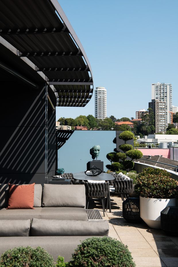 A sophisticated urban rooftop terrace with a large outdoor sectional sofa, dining table and chairs, sculptural topiary planting, and a sculptural bust. The terrace features dark architectural screens and a deep projecting louvred canopy, with a city skyline visible beyond.