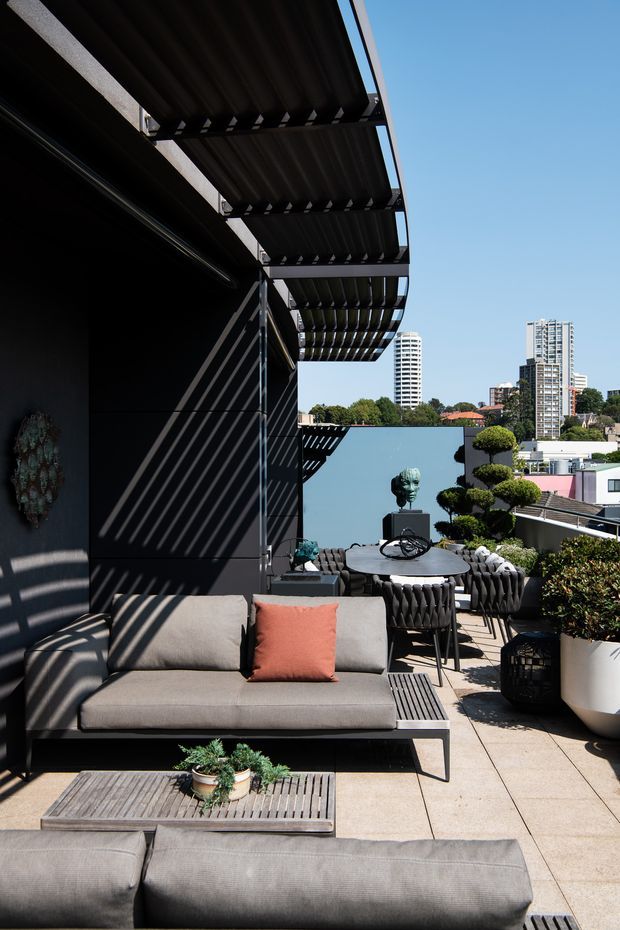 A sophisticated rooftop terrace or elevated outdoor living area overlooking a body of water and urban high-rise buildings. The terrace features a grey outdoor sofa with a coral cushion, a slatted timber coffee table, a round dining table with sculptural dark woven chairs, potted topiary and other plants, and a dark metal pergola or louvred overhead structure casting strong linear shadows. The palette is charcoal, grey, warm timber, greenery, and terracotta, with a bright contemporary atmosphere.