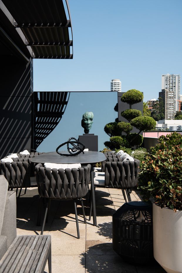 A sophisticated rooftop terrace with a round outdoor dining table and sculptural woven dark-grey chairs. Black architectural screens and pergola-like overhead fins cast strong linear shadows, while manicured topiary, large planters, and a sculptural bust decorate the space. The terrace overlooks a dense urban skyline under a clear blue sky.