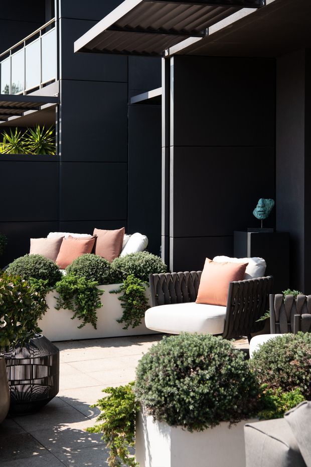 A sophisticated outdoor terrace courtyard with black panelled walls, projecting metal awnings, and a small upper-level balcony. The paved space is furnished with pale upholstered lounge chairs and sofas accented by blush cushions, surrounded by lush clipped shrubs and trailing greenery in large white planters. Strong sunlight and deep shadows create a refined, contemporary atmosphere.