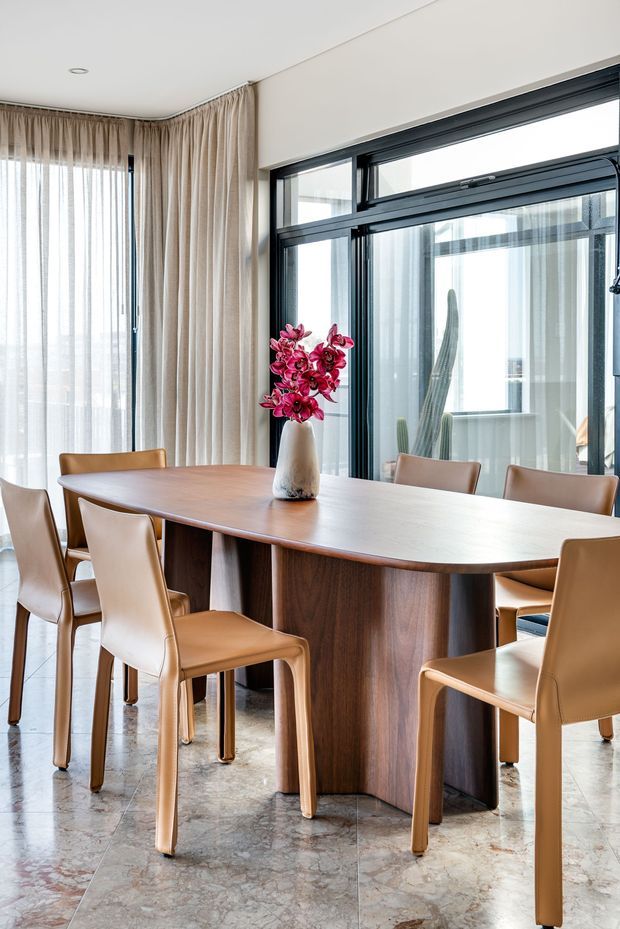 A refined dining area with a large rounded rectangular dark timber dining table surrounded by tan leather dining chairs. Full-height sheer and beige curtains frame expansive black-framed glazing, with a marble or stone-look floor and a vase of pink flowers as a centrepiece. The room is bright, elegant and contemporary, with an adjoining glazed space visible beyond the windows.
