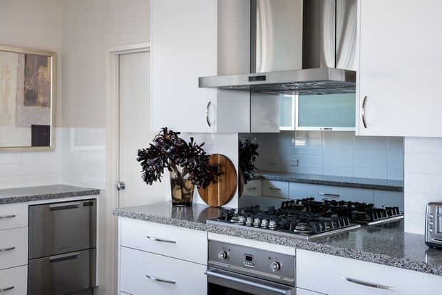 Bright contemporary kitchen with white flat-panel cabinetry, a speckled grey stone benchtop, white horizontal subway-tile splashback, stainless-steel rangehood and built-in oven. The kitchen includes a gas cooktop, integrated dishwasher, toaster and timber serving boards, with a reflective splashback section behind the cooktop.