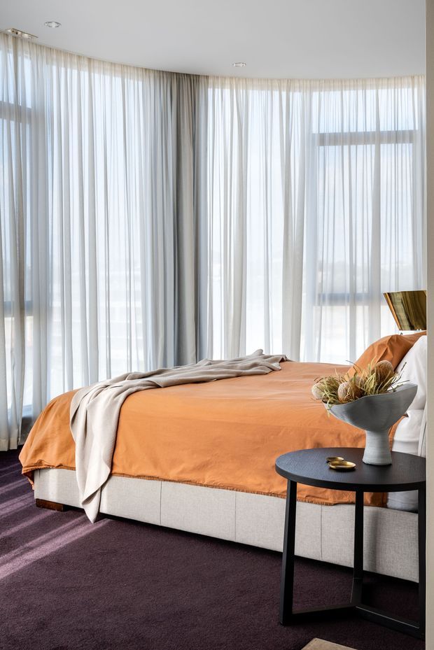 A bright, contemporary bedroom with floor-to-ceiling sheer curtains wrapping around large windows, an upholstered light bed base, and a vivid rust-orange bedspread. A dark round side table with a sculptural vase sits beside the bed, while a deep plum carpet and neutral textiles add contrast. The room has a curved window wall and recessed ceiling lights.