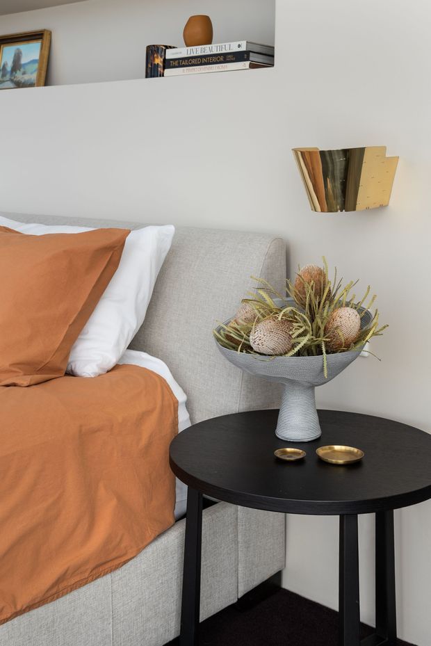 A close-up of a stylish bedroom corner featuring a light grey upholstered bed with rust-orange bedding, a round black bedside table, a sculptural vase of dried botanical arrangements, and a brass wall sconce. A floating shelf displays books and a small ceramic vessel, with framed artwork partially visible.