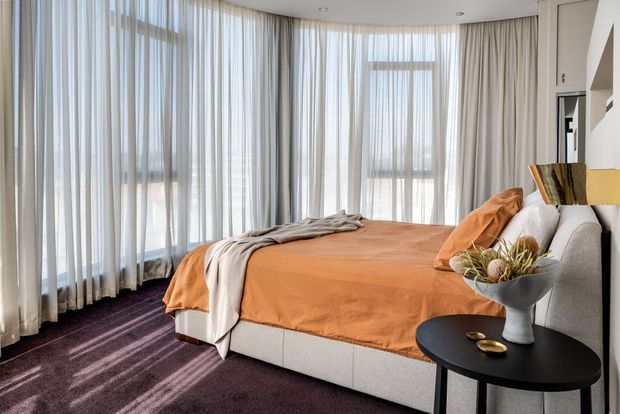 Bright contemporary bedroom with a large upholstered bed dressed in an orange bedspread, grey and cream layered throws, and matching orange cushions. Floor-to-ceiling sheer curtains wrap around the room, allowing soft daylight onto a dark purple carpet. A small black round side table holds a sculptural vase and decorative objects, while built-in joinery is visible along the right wall.
