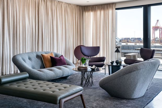 A refined contemporary living room with sculptural upholstered seating, including a curved patterned sofa, two lounge chairs, and a tufted leather ottoman. Floor-to-ceiling cream curtains frame large dark-framed windows overlooking an urban waterfront or port setting. A dark textured rug, glass coffee tables, and subtle decorative accessories create a sophisticated, mid-century-influenced interior.