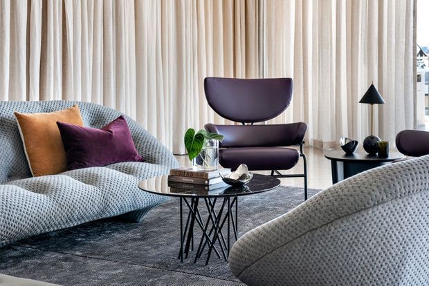 A refined living room with a sculptural upholstered sofa and lounge chair, layered with warm ochre and deep plum cushions. A round glass coffee table with a geometric black metal base sits on a dark textured rug, while full-height cream curtains create a soft backdrop. Black accent lighting and dark furniture provide contrast, and abundant daylight enters from the glazed opening at right.