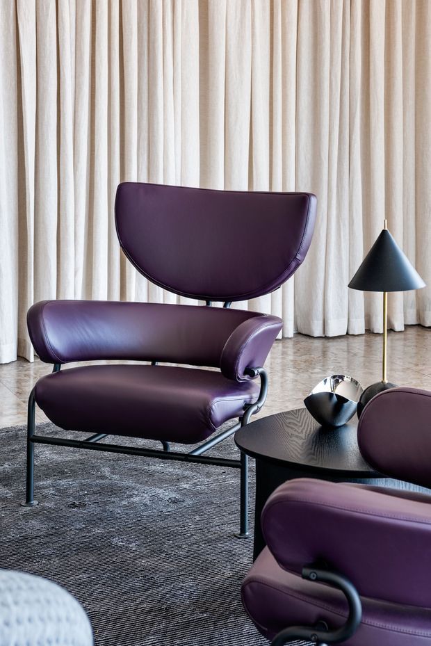 A refined living room vignette featuring sculptural deep-purple leather lounge chairs with black metal frames, a round black coffee table, and a matching floor lamp. Full-height cream curtains form the backdrop, while a textured grey rug and polished stone or concrete-look flooring add layered neutral finishes. The composition has a sophisticated mid-century-inspired feel with strong curved furniture forms and restrained contemporary styling.