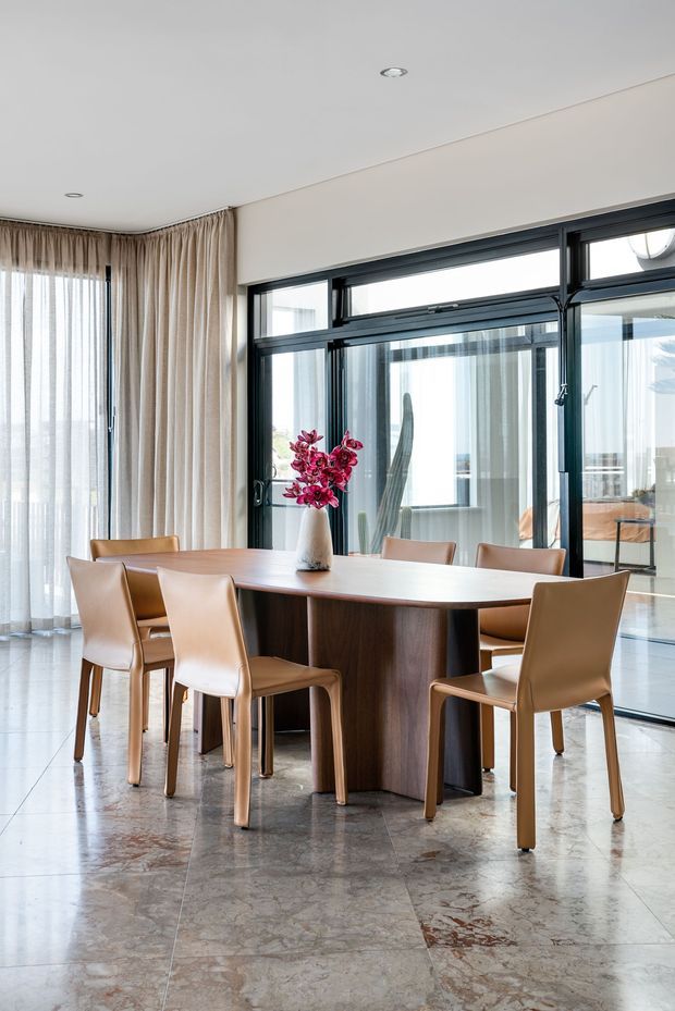 A bright, refined dining room with a large rectangular dark timber dining table surrounded by tan leather dining chairs. Floor-to-ceiling sheer curtains and beige drapes frame expansive black-framed glazing, while polished stone flooring and a simple floral arrangement complete the contemporary interior.