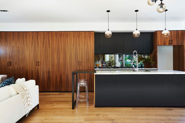 Open-plan contemporary kitchen and living area with extensive warm timber cabinetry and matching timber flooring. A matte black island and upper cabinetry contrast with white benchtops and a mirrored or reflective splashback. Pendant lights hang above the island, while the edge of a white sofa and a small bar-style seating area are visible in the foreground.