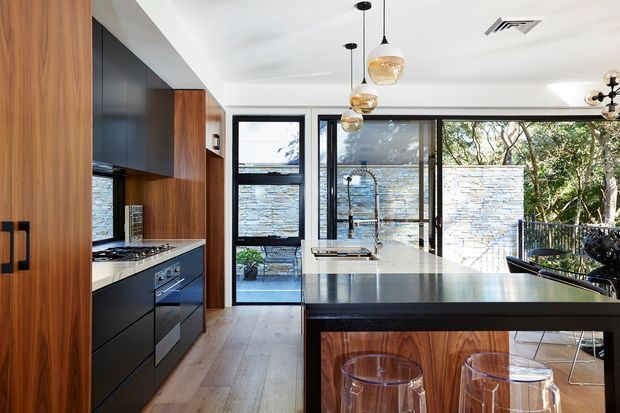 Contemporary kitchen with a large dark stone island, black and timber cabinetry, integrated cooking appliances, pendant lighting and full-height glazing opening toward a stone courtyard wall and trees. The space has a refined, warm material palette with strong indoor-outdoor connection.