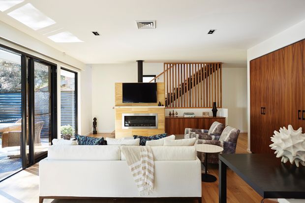 Bright contemporary living room with a large white sectional sofa, timber feature fireplace surround, wall-mounted television, and a wood-slat stair or balustrade screen. Full-height black-framed glazing opens to an outdoor area, while dark built-in cabinetry and a black console add contrast. The room has a light, airy palette with warm timber accents and recessed ceiling lighting.