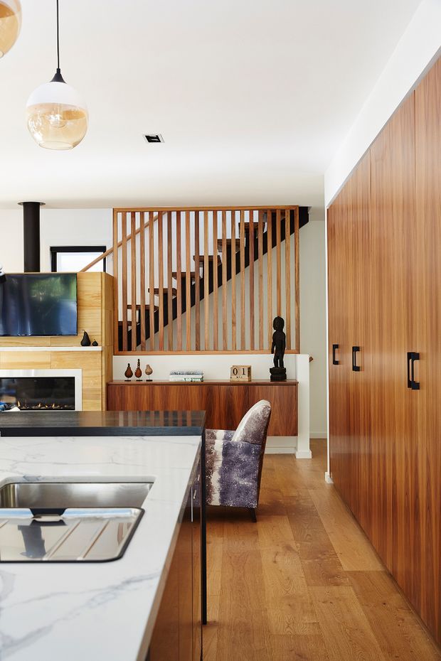 A bright open-plan kitchen and living area with a large white marble-look island, integrated sink, timber cabinetry, and warm timber flooring. A timber slatted screen encloses a staircase, while a wood-burning fireplace, television, low sideboard, sculptural pendant lights, and upholstered dining or bar chair add to the space. The design combines clean contemporary lines with warm mid-century-inspired timber detailing.