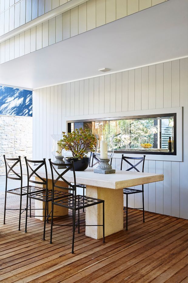 Bright dining area on a timber deck, featuring a substantial light stone or concrete dining table with pedestal base, black metal cross-back chairs, white vertical wall panelling, and a large black-framed mirror. The open, airy space has a calm coastal-contemporary feel with greenery and natural light.