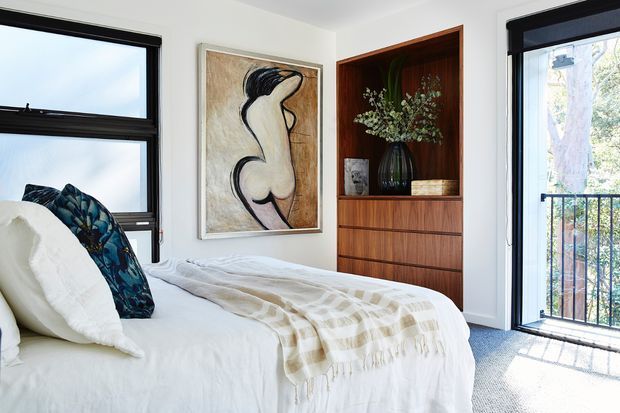 Bright, compact bedroom with a white upholstered bed dressed in white bedding and a beige checked throw. Black-framed windows and a glazed balcony door bring in abundant daylight, while a built-in dark timber display and drawer unit provides storage. The room includes a large neutral-toned figurative artwork and botanical decor, creating a clean, contemporary feel.