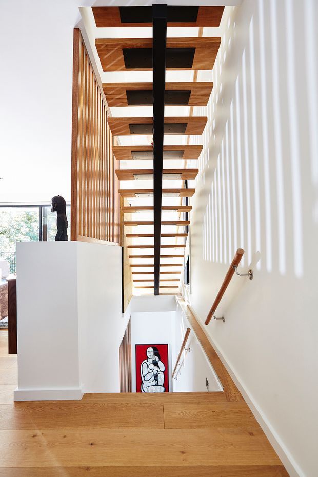 A bright contemporary stairway viewed from the upper landing, featuring warm timber stair treads and handrails, a dramatic open timber slat ceiling with black recessed panels and a central dark beam, white walls, and vertical timber screening. Sunlight creates strong linear shadows on the wall, with artwork visible below and a glimpse of an adjacent living area.