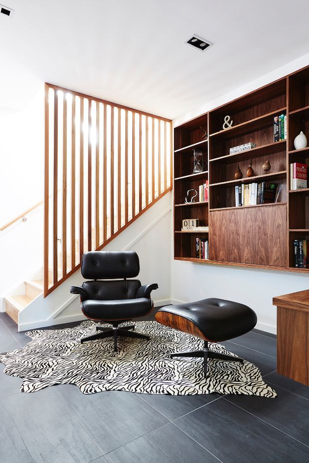 Bright contemporary reading and lounge area beside a staircase, featuring a slatted timber stair balustrade, extensive dark timber built-in shelving, black leather lounge chair and ottoman, and a bold black-and-white patterned rug over dark flooring.