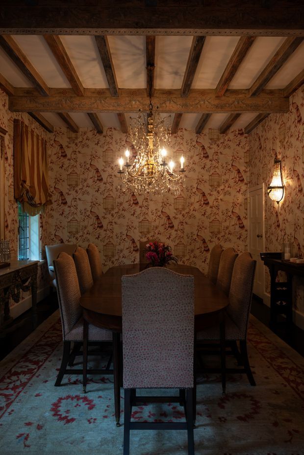 A richly decorated formal dining room with a large oval wooden dining table surrounded by upholstered chairs. The room features ornate patterned wallpaper, exposed dark timber ceiling beams with decorative carved detailing, a large crystal chandelier, a traditional wall sconce, draped window treatments, and a patterned area rug. The lighting is warm and atmospheric, creating a historic, luxurious interior.