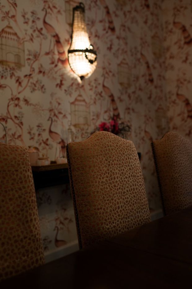 Moody dining room interior with a dark wooden dining table and upholstered dining chairs. The walls feature ornate patterned wallpaper with birds, floral motifs and decorative cages, complemented by a glowing pendant light with a woven or beaded shade. A sideboard with small decor and flowers is visible in the background.