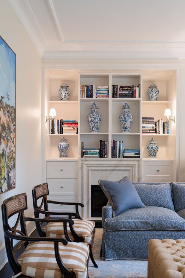 A refined traditional living room with custom white built-in shelving and cabinetry surrounding a marble-trimmed fireplace. The shelves display books and blue-and-white porcelain, while blue patterned upholstery, a tufted ottoman, cane-backed armchairs, and a large framed artwork create a layered, elegant interior.
